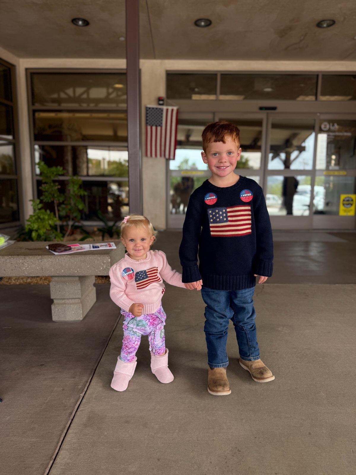 Riley's two children holding hands outside the polling place, wearing American flag sweaters and I Voted stickers